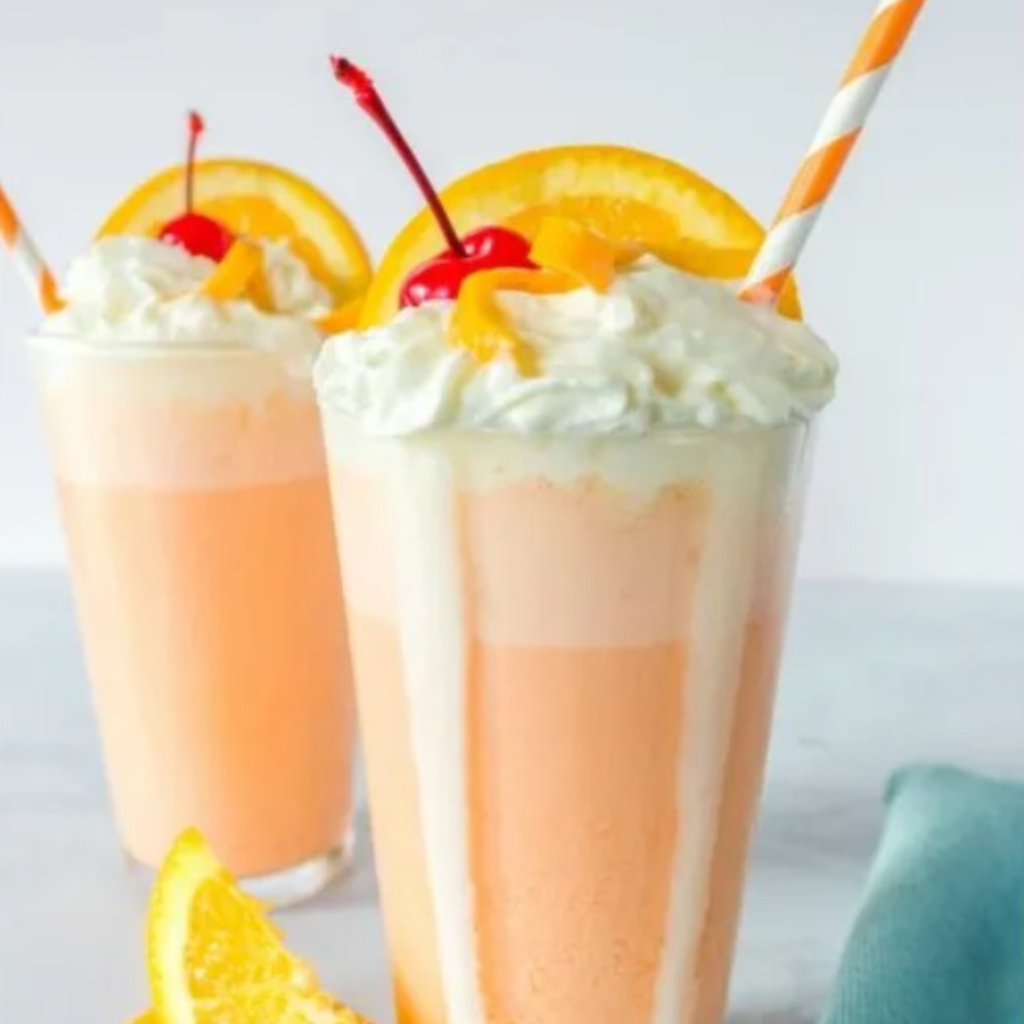 A creamy orange creamsicle milkshake topped with whipped cream, garnished with an orange slice and cherry, served in a tall glass.